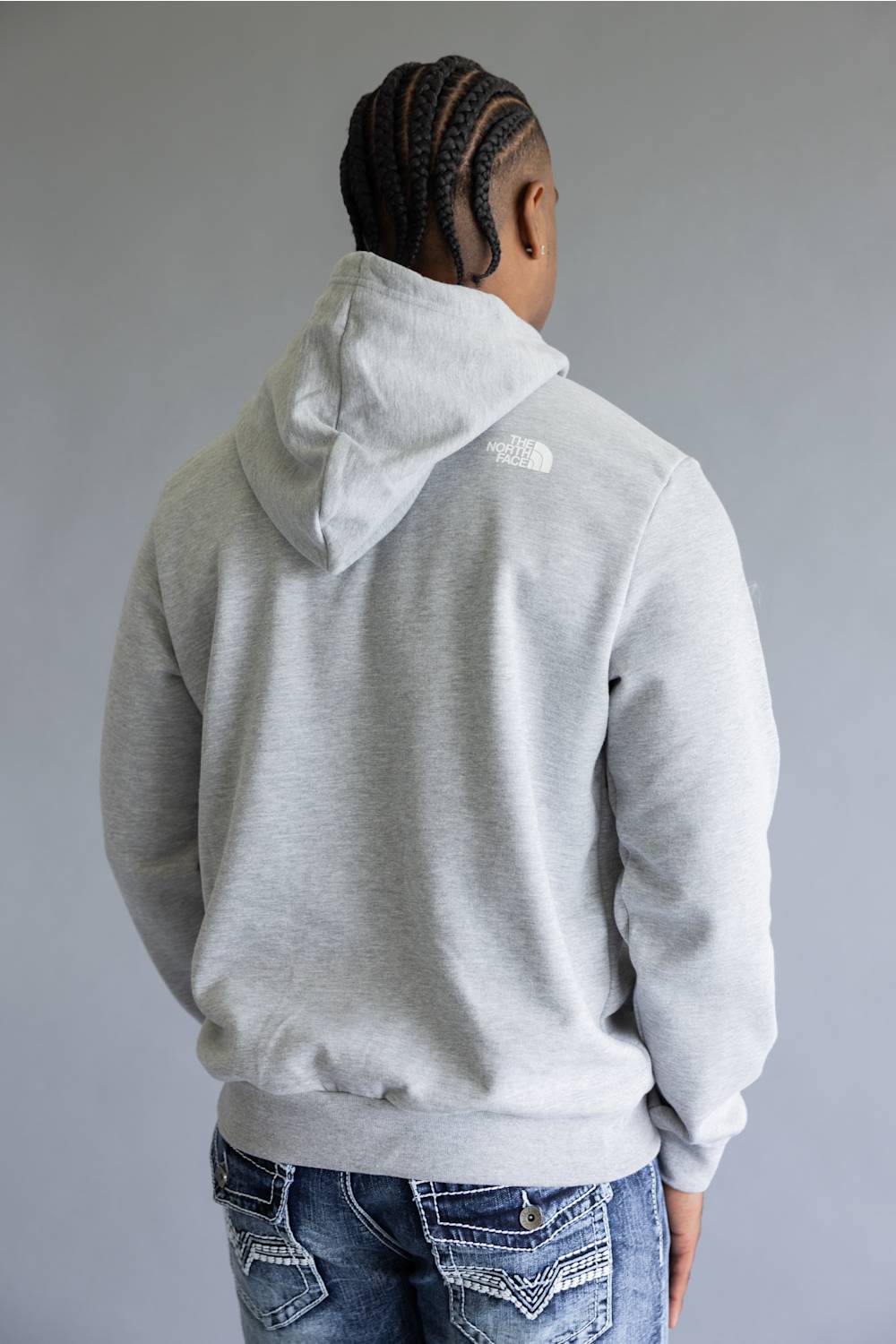A man wearing a light gray heather pullover hoodie with a red 'The North Face' logo patch on the left chest, paired with blue jeans, against a solid gray studio background.