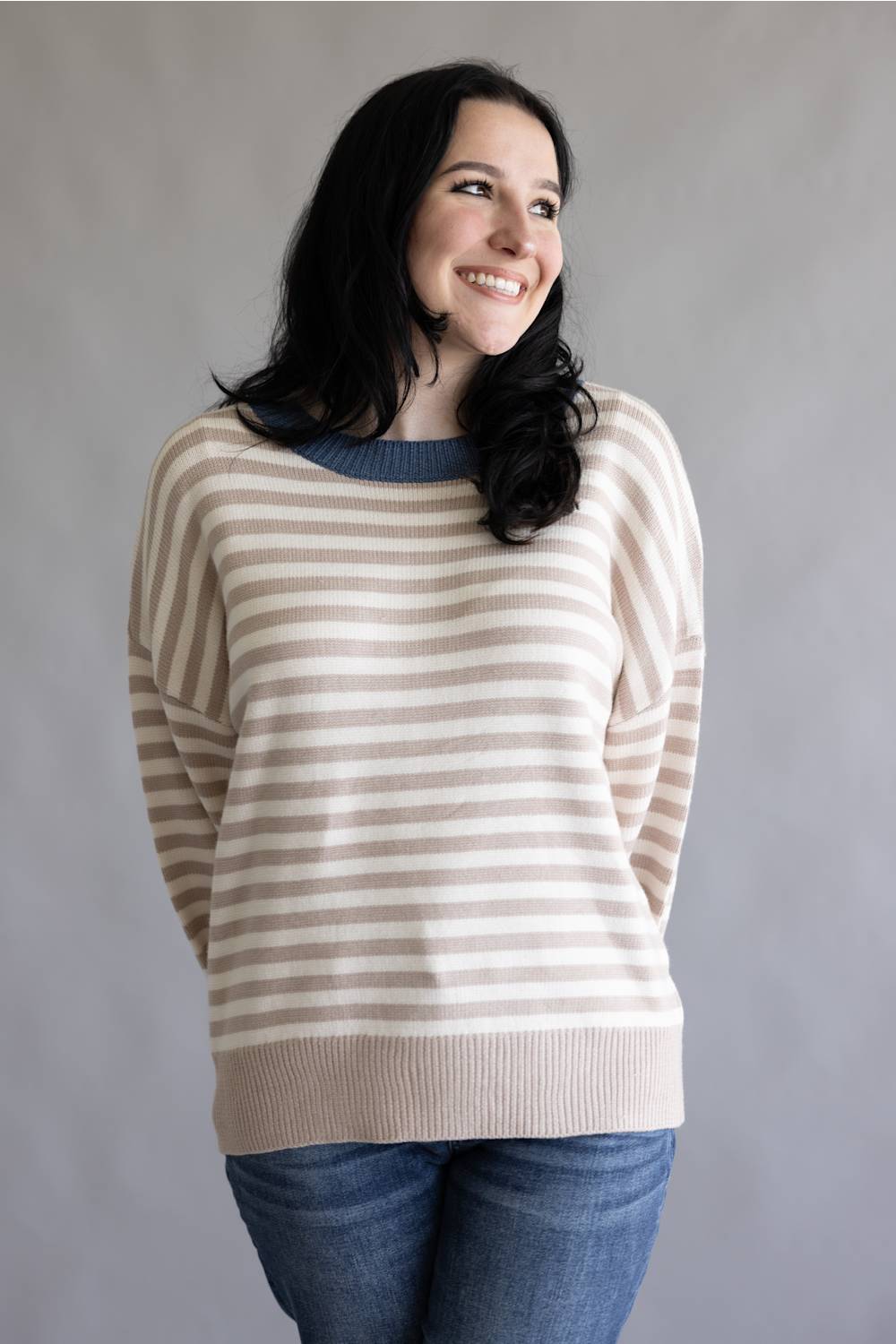 A woman with long black hair smiling and looking to the side, wearing a beige and white horizontally striped knit sweater with a blue crew neck, paired with blue jeans, against a plain gray background.
