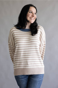 A woman with long black hair smiling and looking to the side, wearing a beige and white horizontally striped knit sweater with a blue crew neck, paired with blue jeans, against a plain gray background.