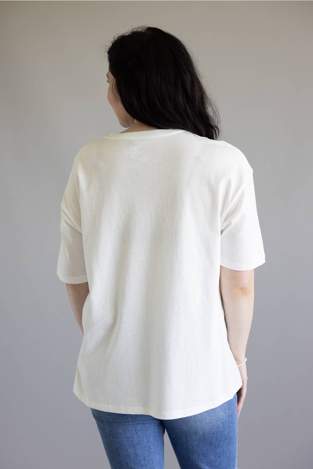 A woman viewed from behind wearing an oversized off-white short-sleeve t-shirt and blue jeans, standing against a plain light gray wall.