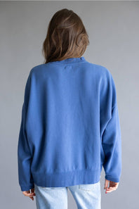 A person shown from the back wearing an oversized blue crewneck sweatshirt and light wash jeans against a plain gray studio background.