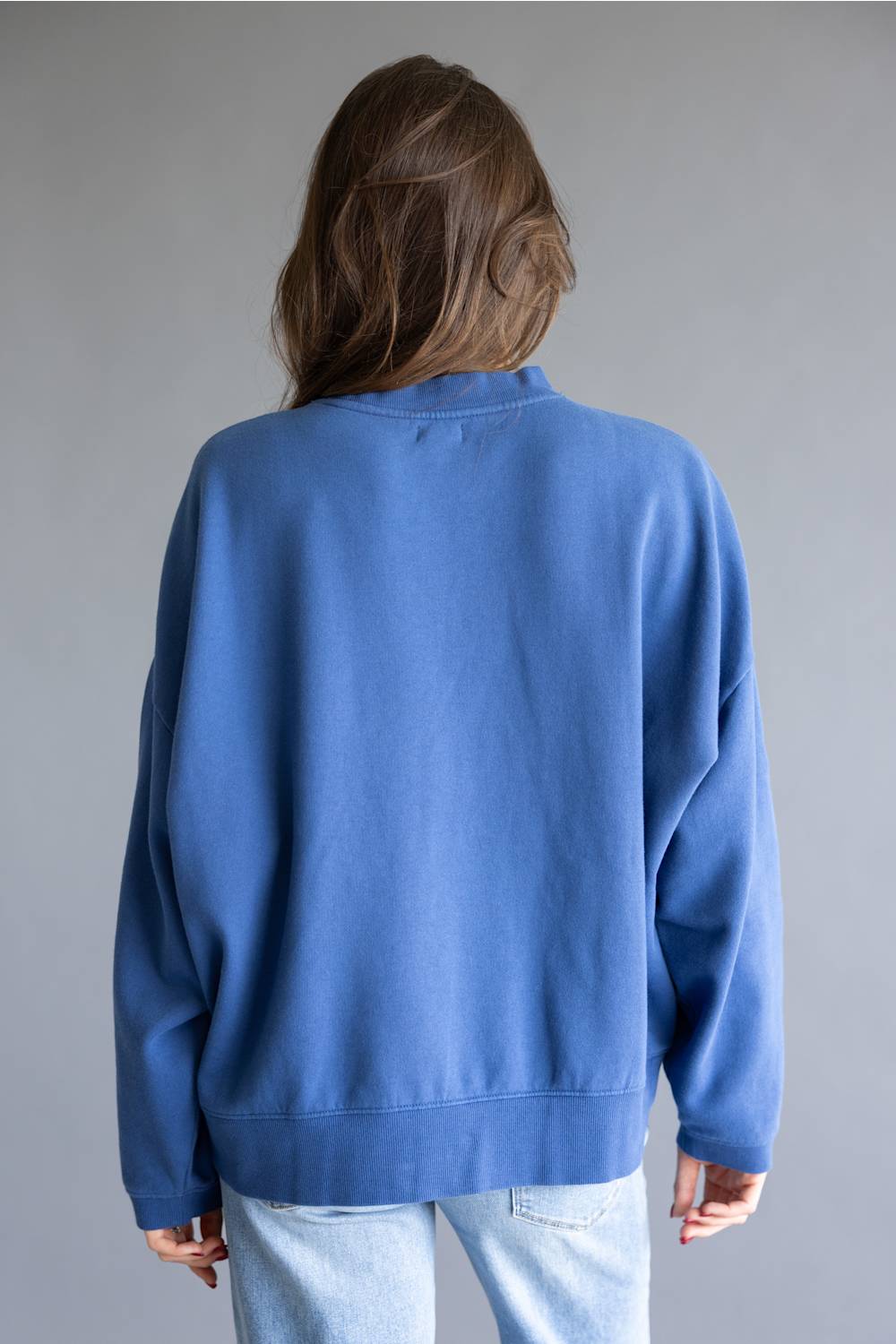 A person shown from the back wearing an oversized blue crewneck sweatshirt and light wash jeans against a plain gray studio background.