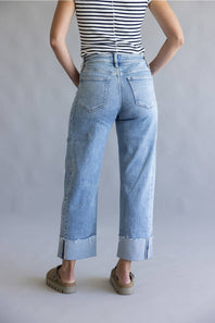 A person shown from the back wearing light-wash cropped jeans with rolled cuffs, a black-and-white striped top, and brown sandals against a plain light gray studio background.