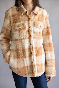 A woman wearing a fuzzy sherpa-style plaid button-up shirt jacket in warm beige and cream tones, paired with dark blue jeans, against a plain gray studio background.