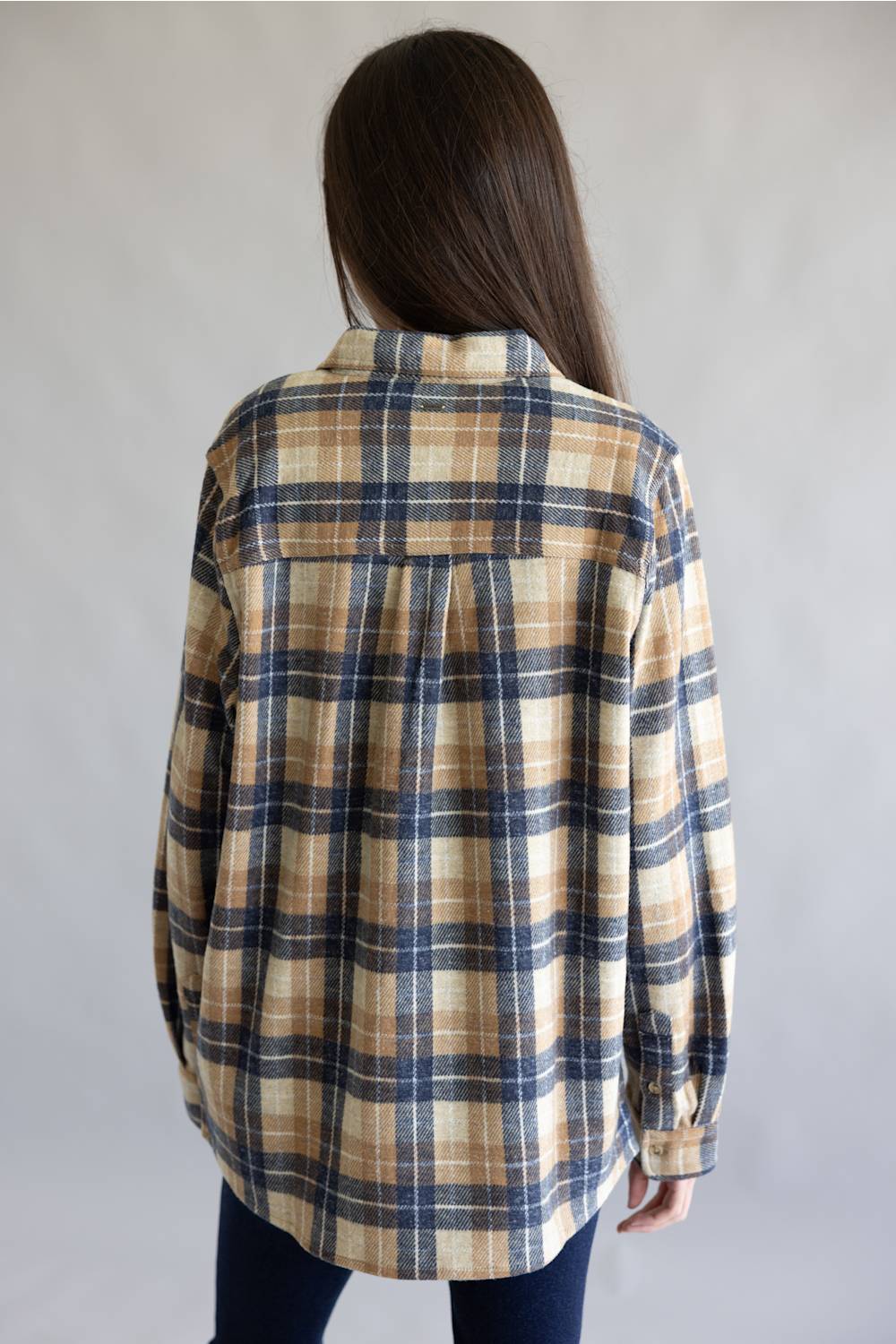 A woman viewed from behind wearing an oversized long-sleeve plaid button-up shirt in beige, navy, and brown tones, paired with dark blue jeans, against a plain light gray background.