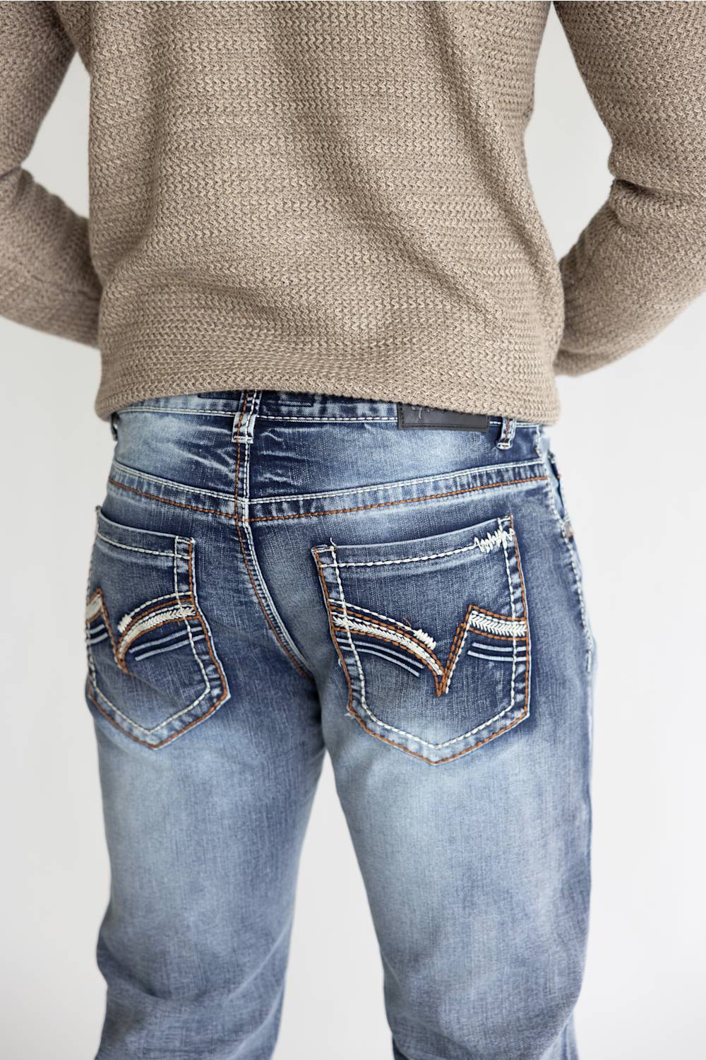 Rear view of a person wearing light blue denim jeans with decorative contrast stitching and embroidery on the back pockets, paired with a beige knit sweater against a plain light background.