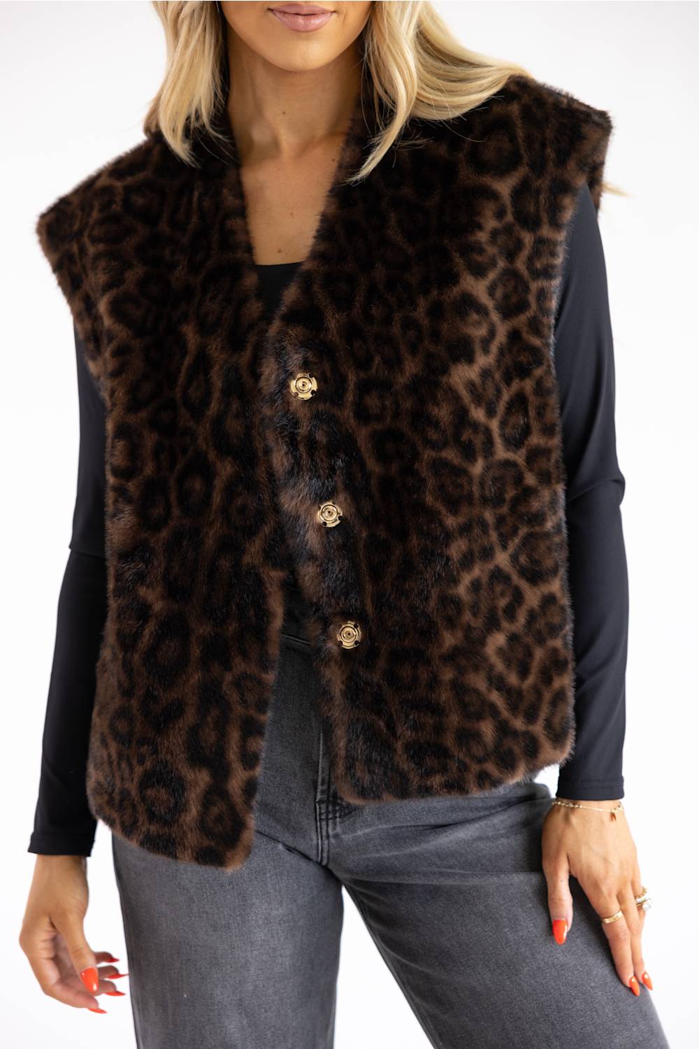 A woman wearing a brown and black leopard-print faux fur vest with gold-tone buttons over a black long-sleeve top and gray jeans.