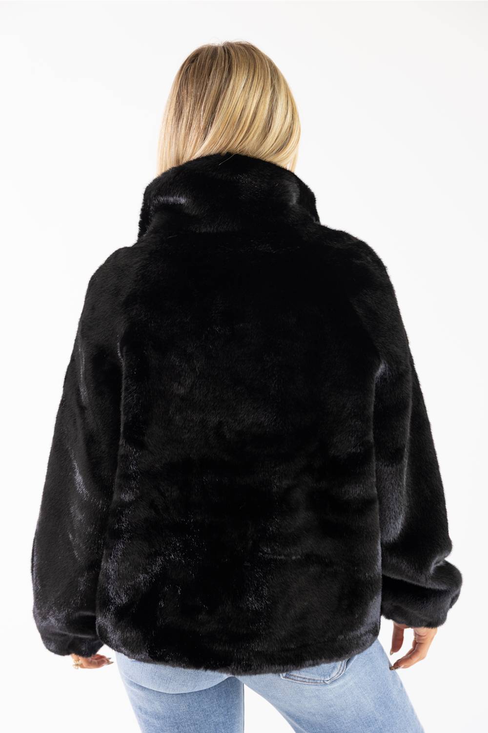 A woman viewed from behind wearing a black faux fur jacket and light blue jeans against a plain white background.