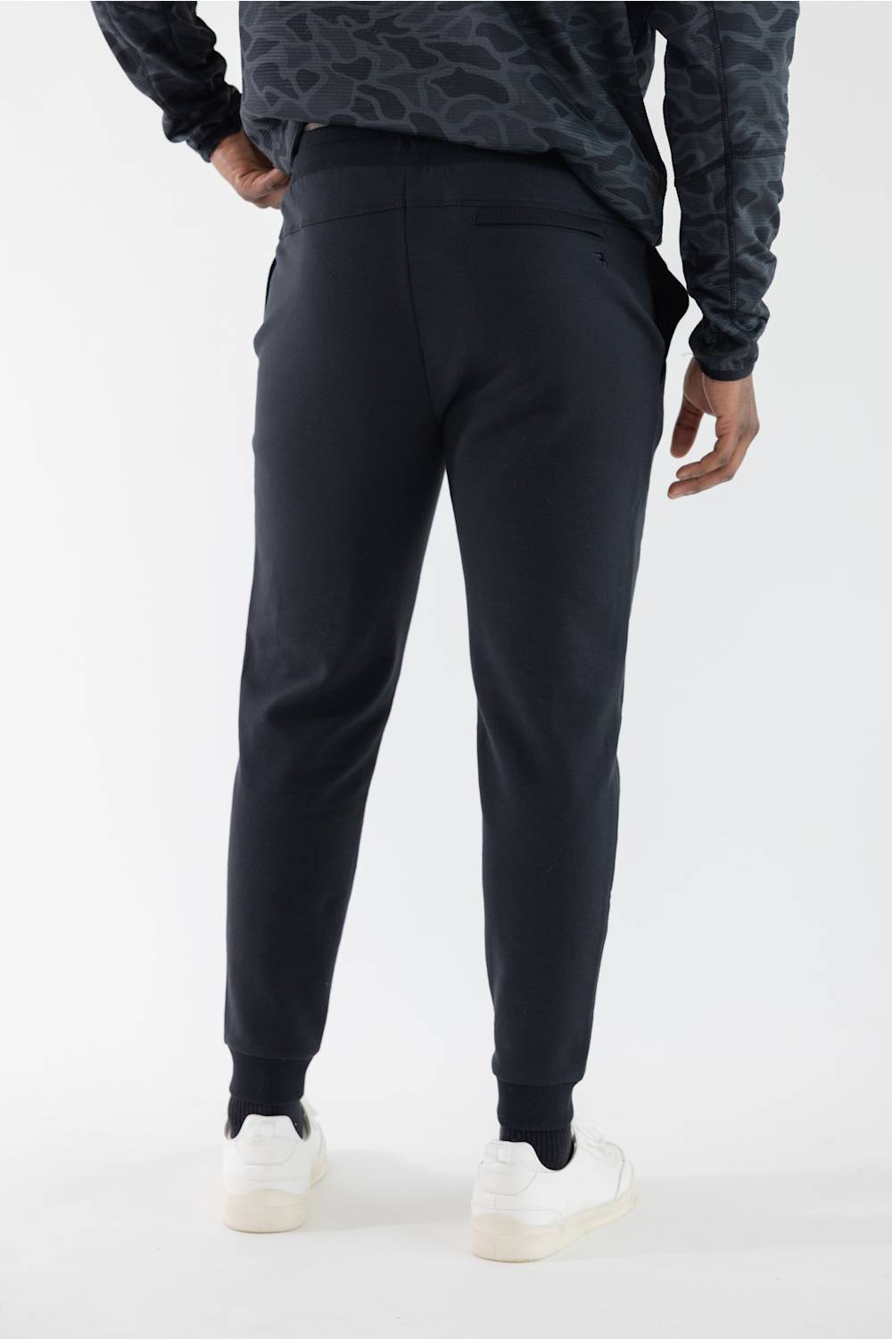 A person shown from the back wearing black jogger-style pants with elastic cuffs, a dark patterned long-sleeve top, and white sneakers against a clean white studio background.
