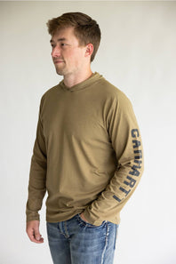 A man wearing a tan long-sleeve hooded shirt with 'Carhartt' printed vertically in black on the right sleeve, paired with blue jeans, photographed against a plain light gray background.