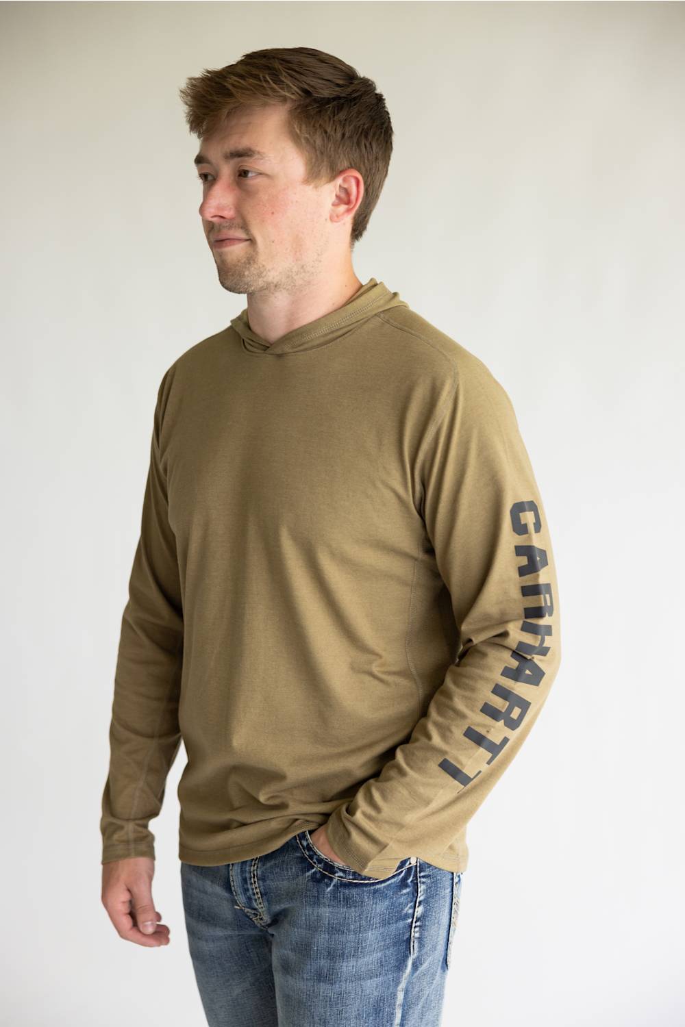 A man wearing a tan long-sleeve hooded shirt with 'Carhartt' printed vertically in black on the right sleeve, paired with blue jeans, photographed against a plain light gray background.