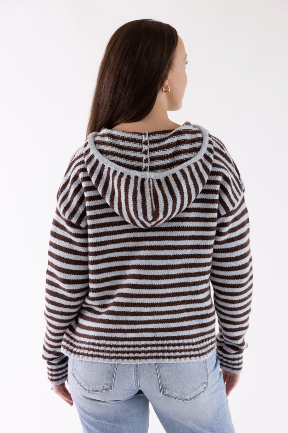 A woman viewed from the back wearing a brown and white horizontally striped knit hoodie with a gray hood trim, paired with light blue jeans, against a plain white background.