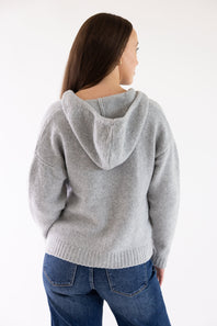 A woman shown from the back wearing a light gray knit hooded sweater and blue jeans against a plain white studio background.