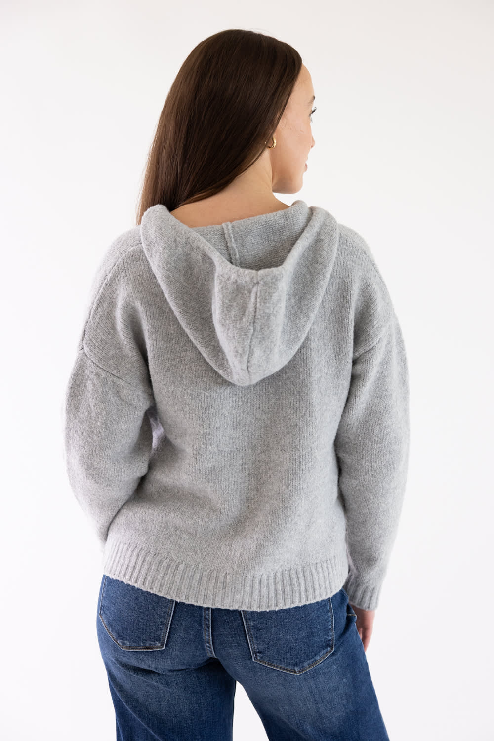 A woman shown from the back wearing a light gray knit hooded sweater and blue jeans against a plain white studio background.