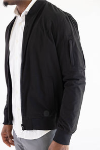 A man wearing a black bomber jacket over a white collared shirt and gray trousers, shown from the chest down against a plain white background.