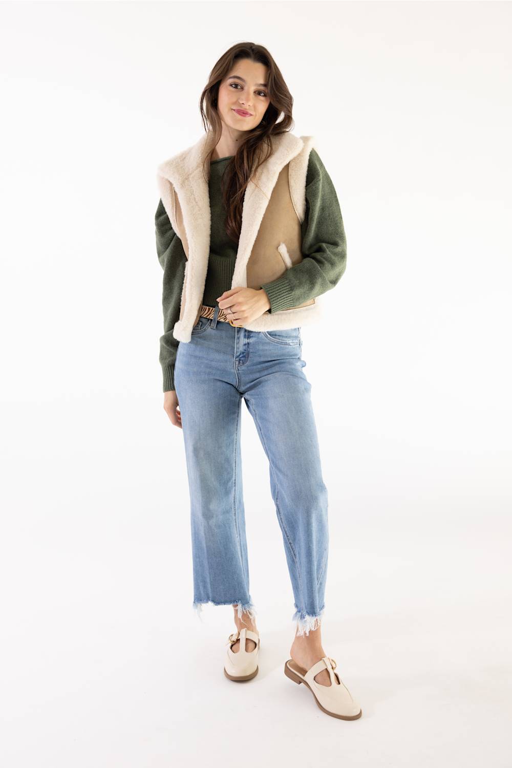 A woman modeling a sleeveless beige shearling-lined jacket with green sleeves, paired with light blue jeans and beige flat shoes, against a plain white background.