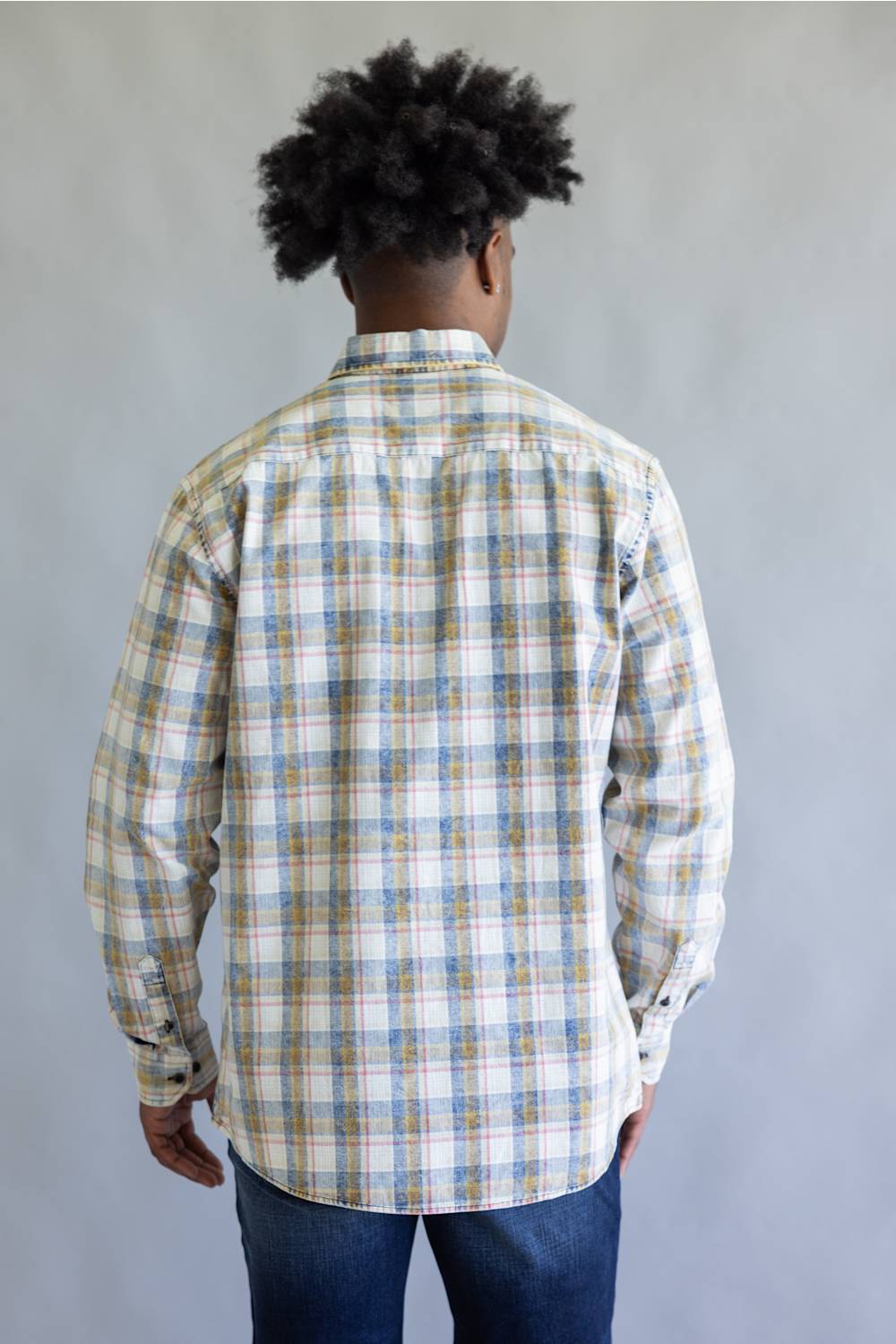 A man viewed from behind wearing a long-sleeve plaid button-up shirt in white, blue, and tan, paired with blue jeans, against a plain gray background.