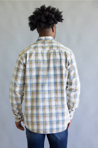 A man viewed from behind wearing a long-sleeve plaid button-up shirt in white, blue, and tan, paired with blue jeans, against a plain gray background.