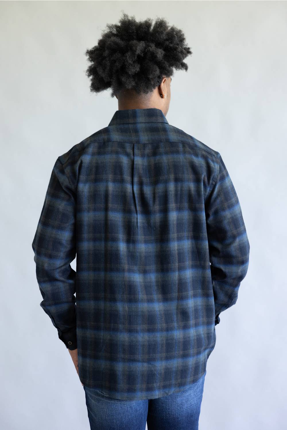A person viewed from the back wearing a dark blue and black plaid button-up shirt with blue jeans, standing against a plain light gray wall.