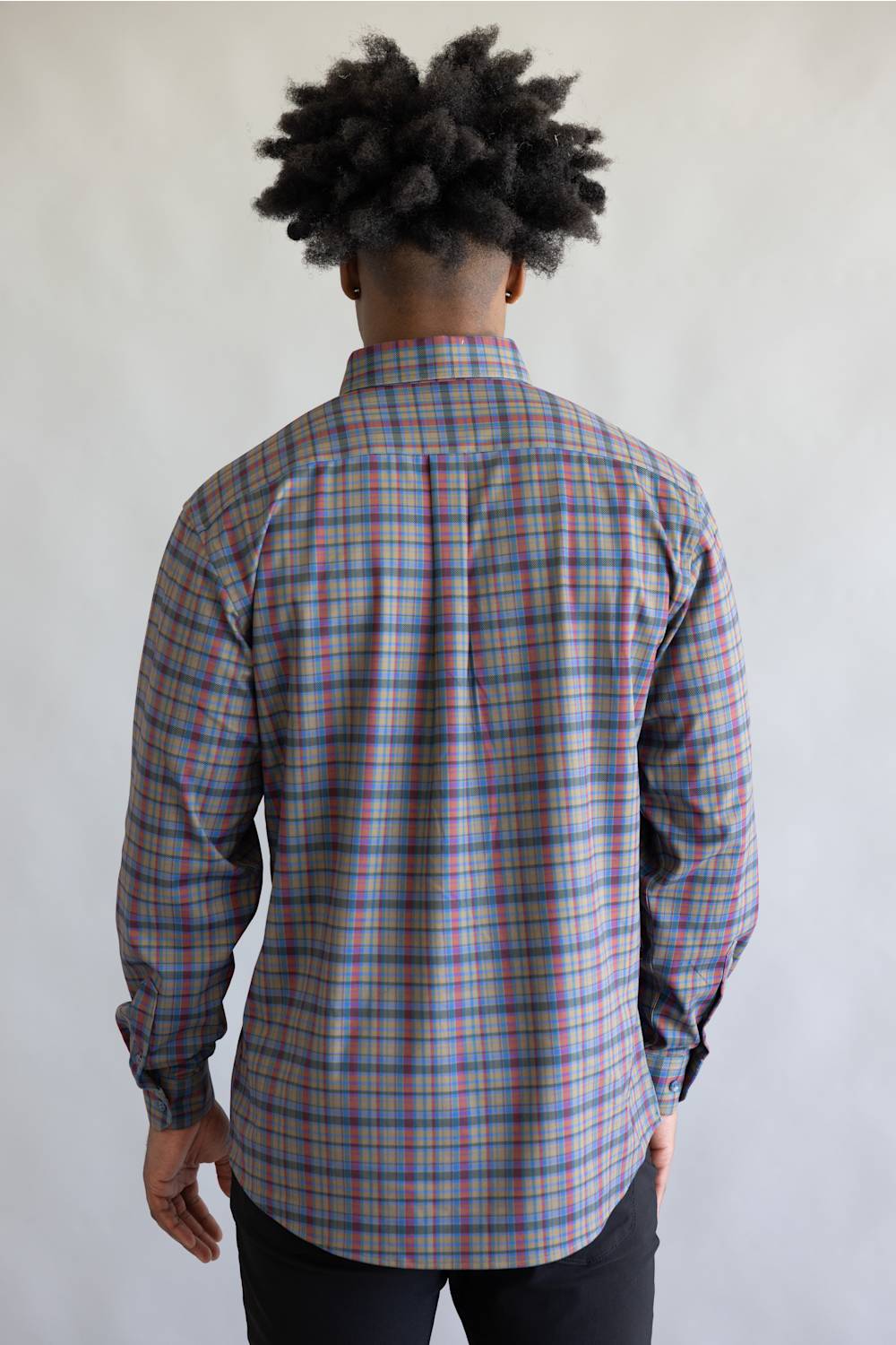 A man viewed from behind wearing a long-sleeve plaid button-down shirt with a blue, red, and beige pattern, paired with black pants, against a plain light gray background.