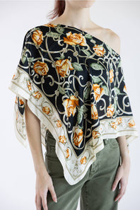 A model wearing a black silk scarf with a yellow rose and green vine pattern, styled as an off-the-shoulder top over olive green pants.