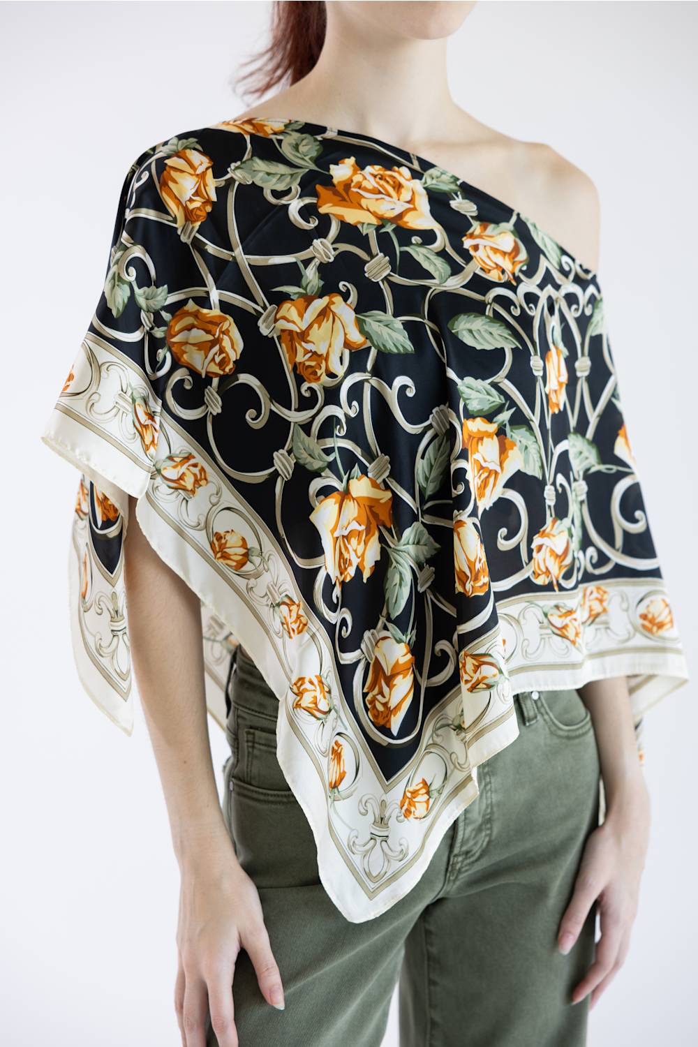 A model wearing a black silk scarf with a yellow rose and green vine pattern, styled as an off-the-shoulder top over olive green pants.