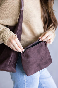 A person wearing a beige ribbed sweater and light blue jeans holds an open dark plum-brown suede crossbody bag, showing the interior pocket. The background is a clean, neutral gray studio backdrop.