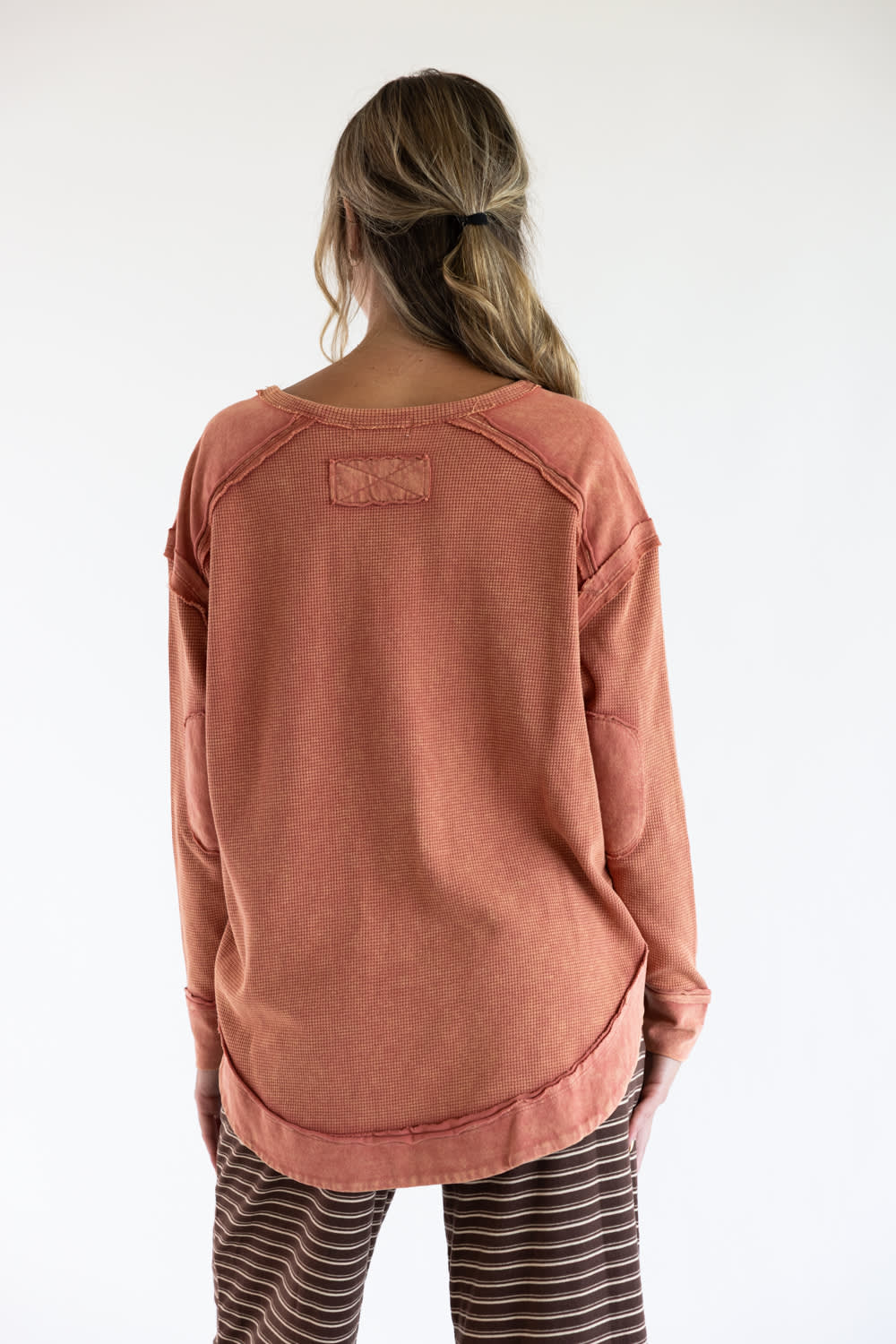 A woman viewed from behind wearing a dusty rose long-sleeve top and brown-and-white striped pants against a plain white background.