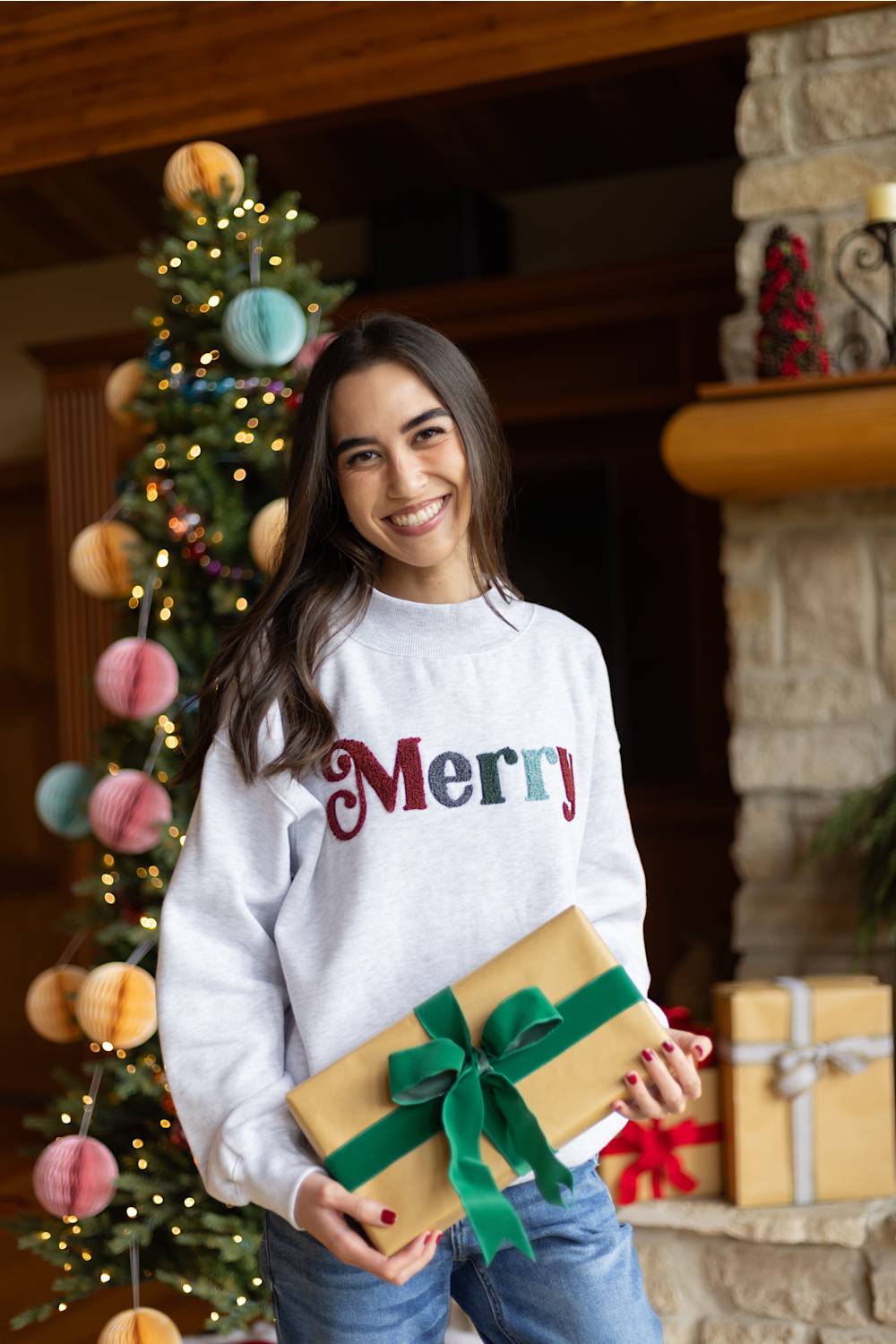 A smiling woman in a white sweatshirt with 'Merry' written across the chest holds a wrapped gift box with a green ribbon. She is standing indoors in front of a decorated Christmas tree and stone fireplace.