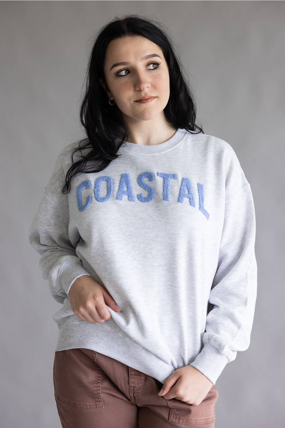 A woman wearing a light gray heather crewneck sweatshirt with 'COASTAL' printed in blue arched letters across the chest, paired with brown pants, against a plain gray studio background.