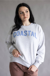 A woman wearing a light gray heather crewneck sweatshirt with 'COASTAL' printed in blue arched letters across the chest, paired with brown pants, against a plain gray studio background.