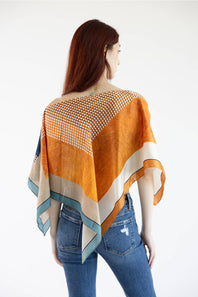 A woman shown from the back wearing a loose, draped orange and beige patterned wrap or poncho over blue jeans against a plain white studio background.