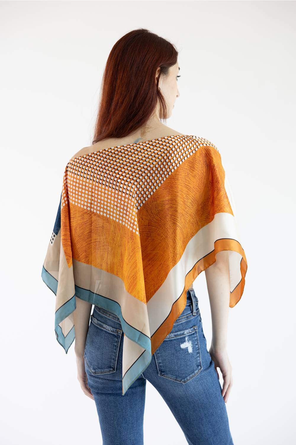 A woman shown from the back wearing a loose, draped orange and beige patterned wrap or poncho over blue jeans against a plain white studio background.