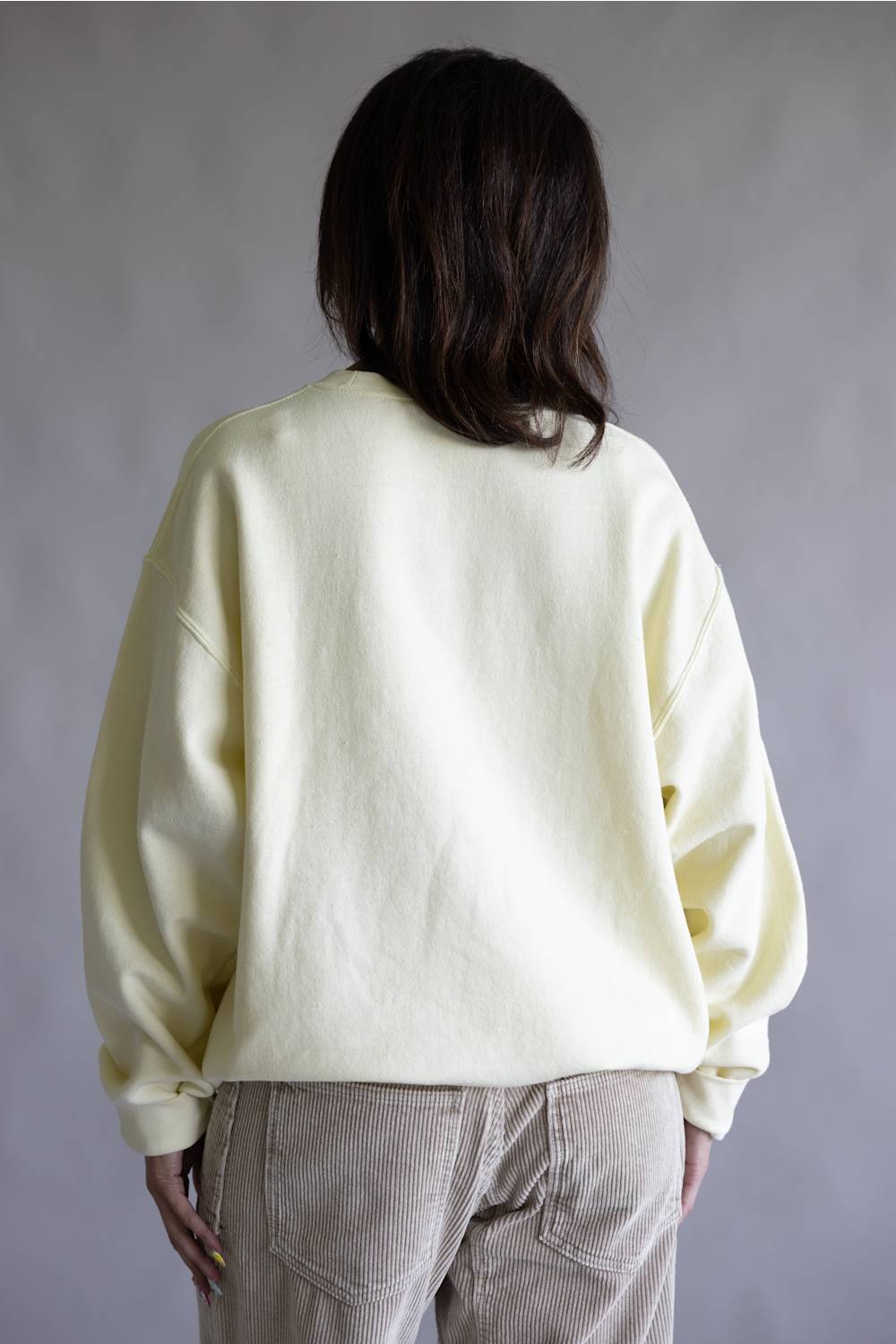 A person shown from the back wearing a pale cream-colored oversized crewneck sweatshirt and light beige corduroy pants against a neutral gray background.