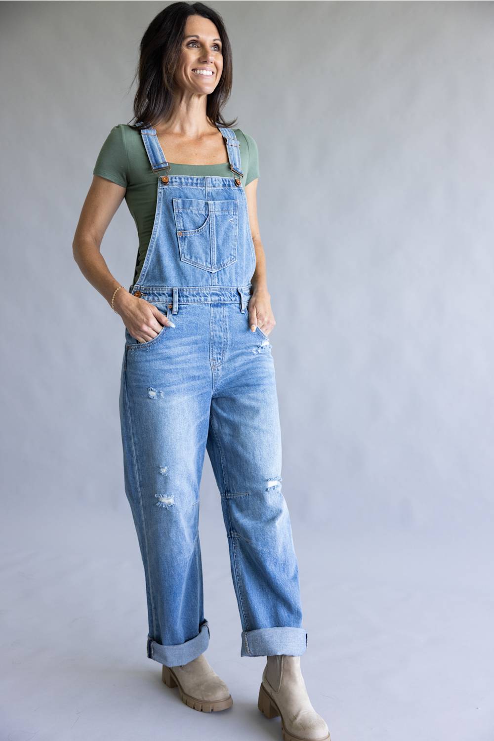 A woman modeling light-wash blue denim overalls with distressed details, cuffed hems, and adjustable bib straps, worn over a sage green t-shirt and beige ankle boots, against a neutral gray studio backdrop.