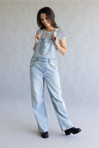 A woman modeling light-wash denim overalls over a black and white striped t-shirt, standing against a neutral gray studio backdrop.