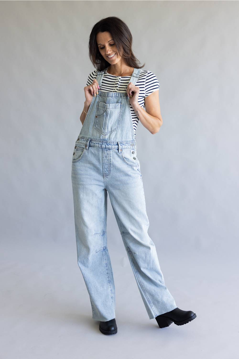 A woman modeling light-wash denim overalls over a black and white striped t-shirt, standing against a neutral gray studio backdrop.