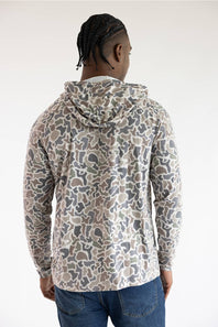 A man viewed from behind wearing a hooded long-sleeve shirt with an abstract camouflage pattern in beige, gray, and brown, paired with blue jeans against a plain white studio background.