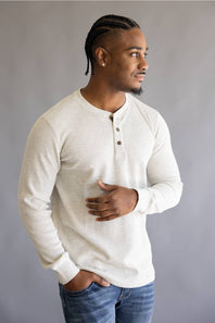 A man wearing a light beige long-sleeve henley shirt and blue jeans, standing against a neutral gray background.
