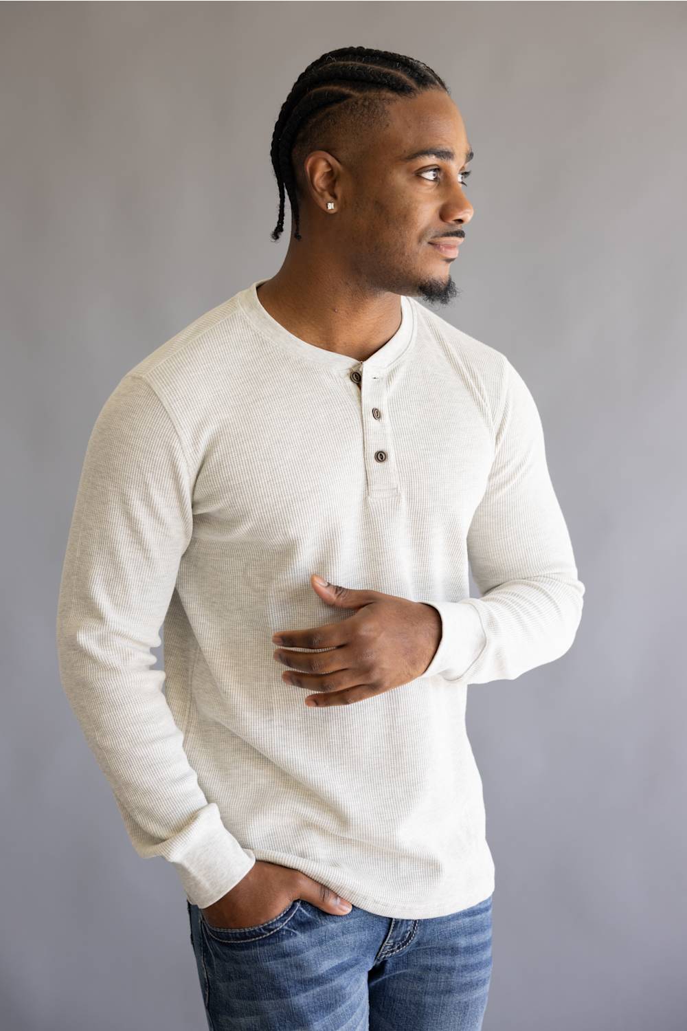 A man wearing a light beige long-sleeve henley shirt and blue jeans, standing against a neutral gray background.