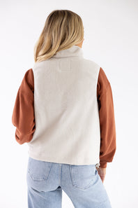 A woman shown from the back wearing a cream fleece vest with contrasting rust-brown sleeves, paired with light blue jeans, against a plain white studio background.