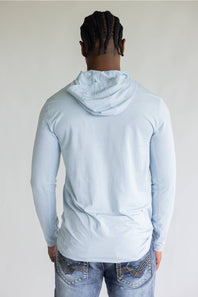 A man viewed from behind wearing a light blue long-sleeve hooded shirt and blue jeans against a plain light gray background.