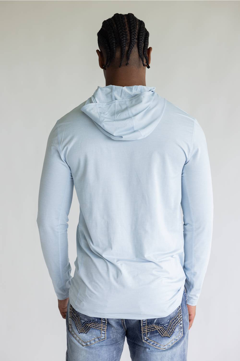 A man viewed from behind wearing a light blue long-sleeve hooded shirt and blue jeans against a plain light gray background.