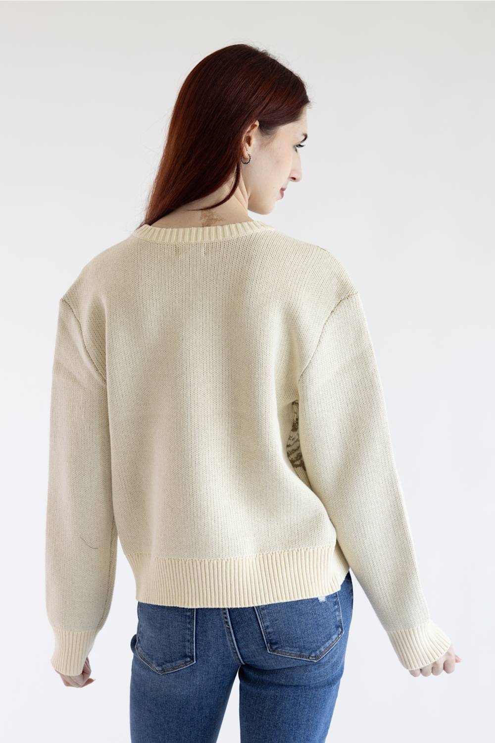 A woman with long auburn hair is shown from the back wearing a cream-colored ribbed knit sweater and blue jeans against a plain white studio background.