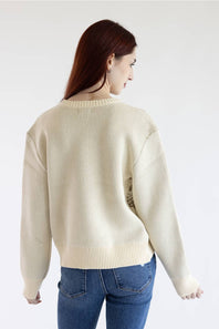 A woman with long auburn hair is shown from the back wearing a cream-colored ribbed knit sweater and blue jeans against a plain white studio background.