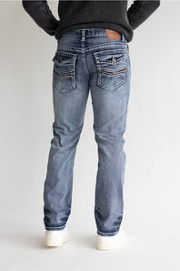 Rear view of a person wearing light blue denim jeans with a black ribbed sweater and white sneakers, standing against a plain light gray studio background.