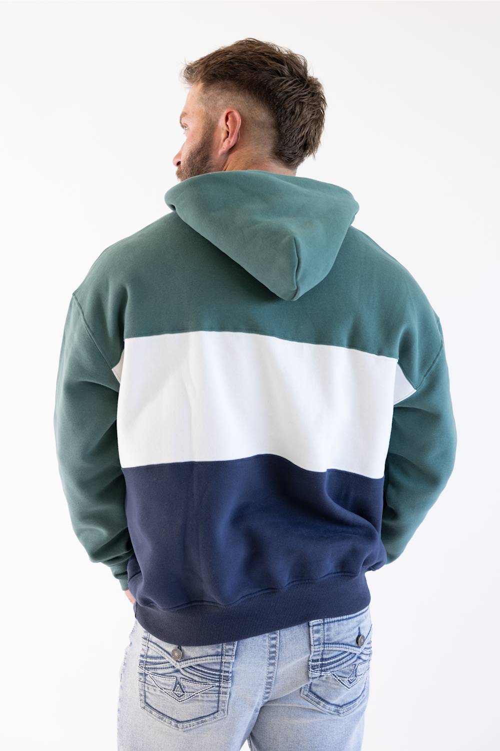 A man viewed from behind wearing a color-block hoodie with a teal green top section, a wide white middle stripe, and a navy blue bottom section, paired with light-wash jeans against a plain white background.
