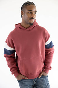 A man wearing a dusty rose red pullover hoodie with navy blue and white stripes on the sleeves, paired with blue jeans, against a plain white background.
