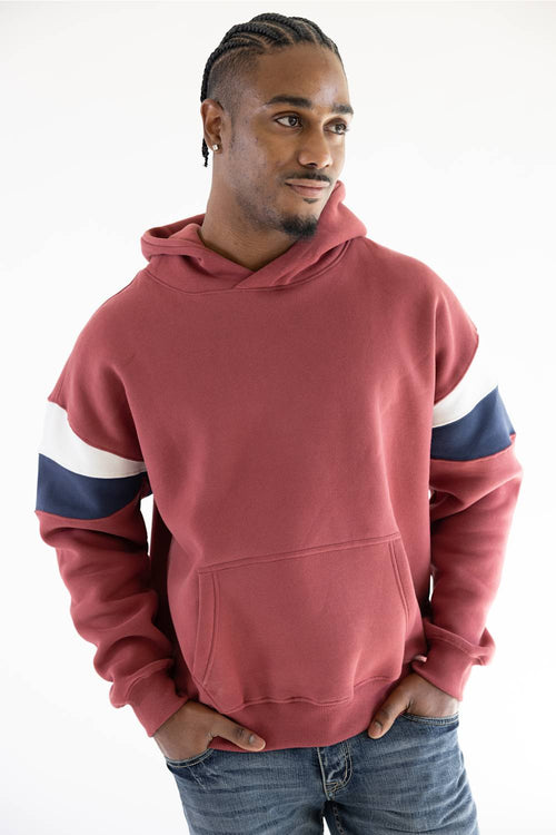 A man wearing a dusty rose red pullover hoodie with navy blue and white stripes on the sleeves, paired with blue jeans, against a plain white background.