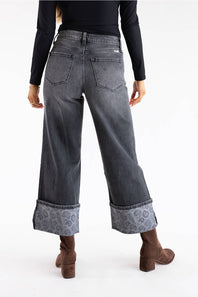 A person shown from the back wearing dark gray wide-leg cropped jeans with a paisley-patterned cuff, a black long-sleeve top, and brown ankle boots against a plain white studio background.